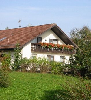 Haus mit rotem Satteldach und Balkon mit roten Blumen, umgeben von grünen Büschen und Wiese | © Ferienhof Schwab