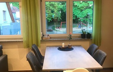 Esszimmer mit grauem Tisch, vier schwarzen Stühlen und einem weißen Hochstuhl vor zwei Fenstern mit grünen Vorhängen, Blick auf einen Spielplatz