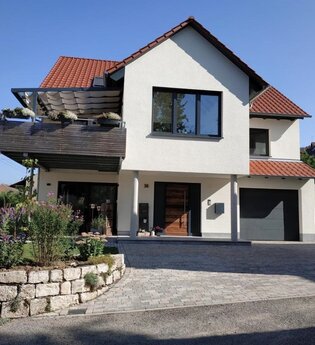 Modernes zweistöckiges Haus mit weißer Fassade, rotem Ziegeldach, Balkon mit Pflanzen und gepflasterter Einfahrt | © Simone Bürckert