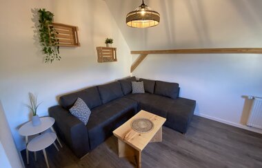 Wohnzimmer mit dunkelgrauem Ecksofa, kleinem Holztisch, zwei weißen Beistelltischen mit Pflanze, hängender Lampe und Holzregalen an weißer Wand | © Ferienwohnung Etzl