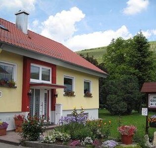 Gelbes Haus mit rotem Dach und Blumenkästen an den Fenstern, davor ein Garten mit bunten Blumen und ein Schild für moderne Gästezimmer mit Dusche und WC | © Gästehaus Gundling