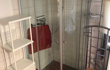 Kleines Badezimmer mit abgerundeter Duschkabine, weißem Regal, rotem Badvorleger und Holzstuhl | © Gästehaus Sabine