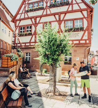 Ein Platz vor einem großen Fachwerkhaus. Auf der rechten Seite steht ein Paar und schaut in eine Karte. Auf der linken Seite stehen zwei Bänke auf welchen Personen sitzen. Walldürn im Odenwald. | © Touristikgemeinschaft Odenwald e.V.