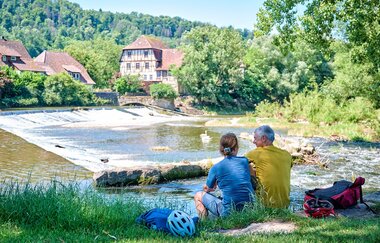 Malerisches Kocherwehr in Forchtenberg, im Vordergrund sitzt ein Paar | © Touristikgemeinschaft Hohenlohe e. V. | Florian Trykowski