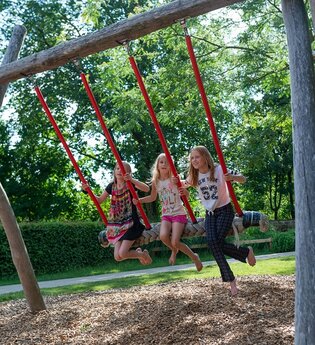 Drei Kinder schaukeln auf einer großen Mehrfachschaukel unter Bäumen