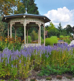 Historischer Pavillon inmitten blühender Stauden unter blauem Himmel.