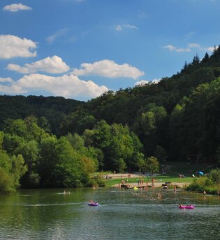 Blick auf den zwischen Wald und Bäumen gelegenen Münster-Badesee. Es sind viele Gäste im Wasser und auf der Liegewiese zu sehen. | © Tourist-Information Creglingen