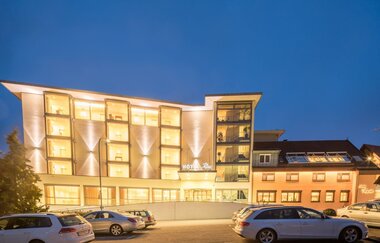 Das Hotel ist nachts hell beleuchtet, davor ist ein Parkplatz für Pkw