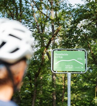 Eine Radfahrerin mit Helm blickt auf ein grünes Schild an der Bannhalde (419 m) im Naturpark Stromberg-Heuchelberg mit der Aufschrift „Spürst Du das auch?“.
