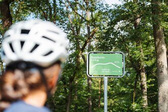 Eine Radfahrerin mit Helm blickt auf ein grünes Schild an der Bannhalde (419 m) im Naturpark Stromberg-Heuchelberg mit der Aufschrift „Spürst Du das auch?“.