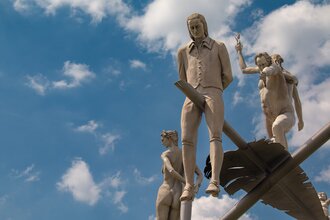 Am Ortseingang von Lauffen am Neckar erhebt sich das eindrucksvolle Kunstwerk „Hölderlin im Kreisverkehr“ des Bildhauers Peter Lenk. Auf einem geschwungenen „H“ balanciert eine überdimensionale Schreibfeder, auf der Friedrich Hölderlin als Kind und als ju | © Stadt Lauffen am Neckar