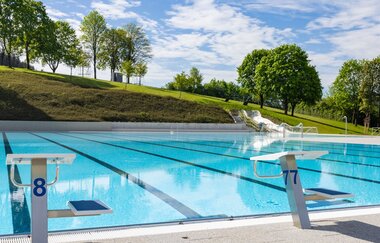 Ein leeres Freibad mit Startblöcken und einer Wasserrutsche im Hintergrund, umgeben von grünen Bäumen und einem blauen Himmel.
