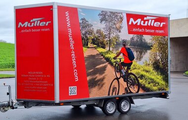 Stehender Fahrradanhänger mit einem Bild von einem Radfahrer am Fluss und das Logo von der Firma Müller Reisen.