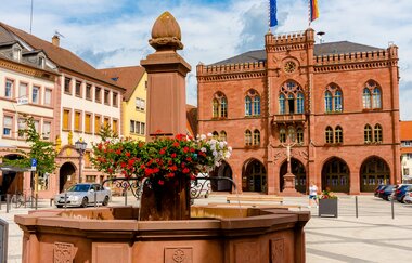 Blick auf den mit roten Geranien geschmückten Brunnen  auf dem Marktplatz in Tauberbischofsheim. Im Hintergrund ist das aus Sandstein gebaute Rathaus zu sehen. | © Tourismusverband "Liebliches Taubertal" e.V.