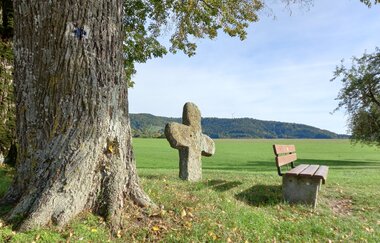 Ein Sühnekreuz und eine Bank stehen unter einem Baum auf einer Wiese mit Hügeln im Hintergrund. | © Petra Natzkowski