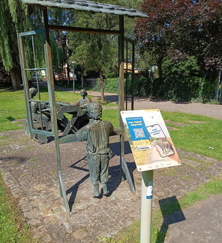 Schild der Weitersehen-Station in Hockenheim vor einer Statue in dem 3 Personen beim Tabakdrehen dargestellt sind