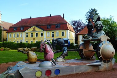Die Skulptur eines sehr modernen und abstrakten Kutscherensembles ist im Vordergrund zu sehen. Im Hintergrund ist das restaurierte und in gelber Farbe erstrahlende Geburtshaus des Revolutionärs Friedrich Heckers sichtbar. | © Landratsamt Rhein-Neckar-Kreis