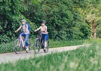 2 Radfahrerinnen fahren auf einem Radweg mit ihren E-Bikes. Der Radweg führt durch Wiesen und Wälder. | © Touristikgemeinschaft Odenwald e.V.