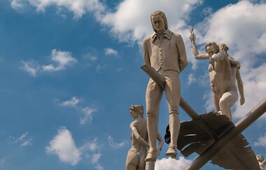 Am Ortseingang von Lauffen am Neckar erhebt sich das eindrucksvolle Kunstwerk „Hölderlin im Kreisverkehr“ des Bildhauers Peter Lenk. Auf einem geschwungenen „H“ balanciert eine überdimensionale Schreibfeder, auf der Friedrich Hölderlin als Kind und als ju | © Stadt Lauffen am Neckar