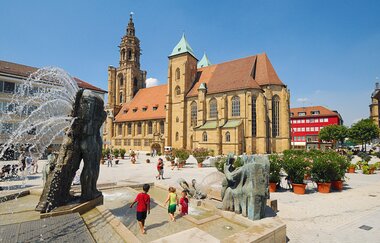 Das Bild zeigt mittig die Kilianskirche davor der, mit hellen Pflastersteinen bebaute Kiliansplatz auf dem mehrere Pflanzkübel mit Oleander zu erkennen sind. Im Vordergrund befindet sich ein begehbarer Springbrunnenmit mehreren Kindern darin | © Heilbronn Marketing GmbH