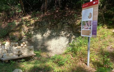 bunte Infotafel am Gaildorfer Geopfad, daneben aufgespapelt kleine, quadratische Sandsteine vor freigelegter Erdschicht