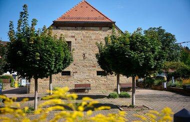 Massives Natursteingebäude mit rotem Satteldach, davor zwei geschnittene Bäume und Sitzbänke – aufgenommen an einem sonnigen Sommertag in Oberderdingen. | © Kraichgau-Stromberg Tourismus e.V.