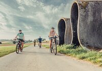 Radler fahren auf einem Fahrradweg, daneben befindet sich eine Skulptur, die eine Straße in Wellen zeigt; im Hintergrund befinden sich Wiesen und Wälder | © Touristikgemeinschaft Odenwald e.V.