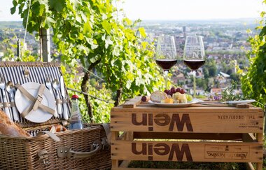Mittig ist eine umgedrehte Weinkiste. Darauf stehen zwei Weingläser mit Rotwein und ein Teller mit Brot,  Käse und Trauben. Daneben steht ein geöffneter Korb, in dem Geschirr zu sehen ist. Im Hintergrund sieht man Weinreben und eine Stadt von oben. | © Heilbronn Marketing GmbH